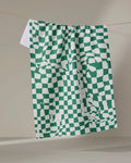 The Verdant kitchen towel by Geometry, featuring a green and white checkered pattern with scattered, abstract squares, hangs on a clothesline against a plain, neutral indoor background.