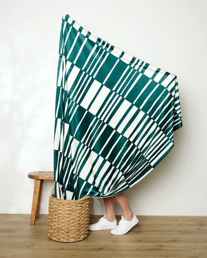 A person stands partly hidden behind Geometry’s Verdelia blanket—green and white striped—draped over a basket, with only their lower legs and white sneakers visible beside a wooden stool on an indoor wood floor.
