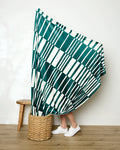 A person stands partly hidden behind Geometry’s Verdelia blanket—green and white striped—draped over a basket, with only their lower legs and white sneakers visible beside a wooden stool on an indoor wood floor.