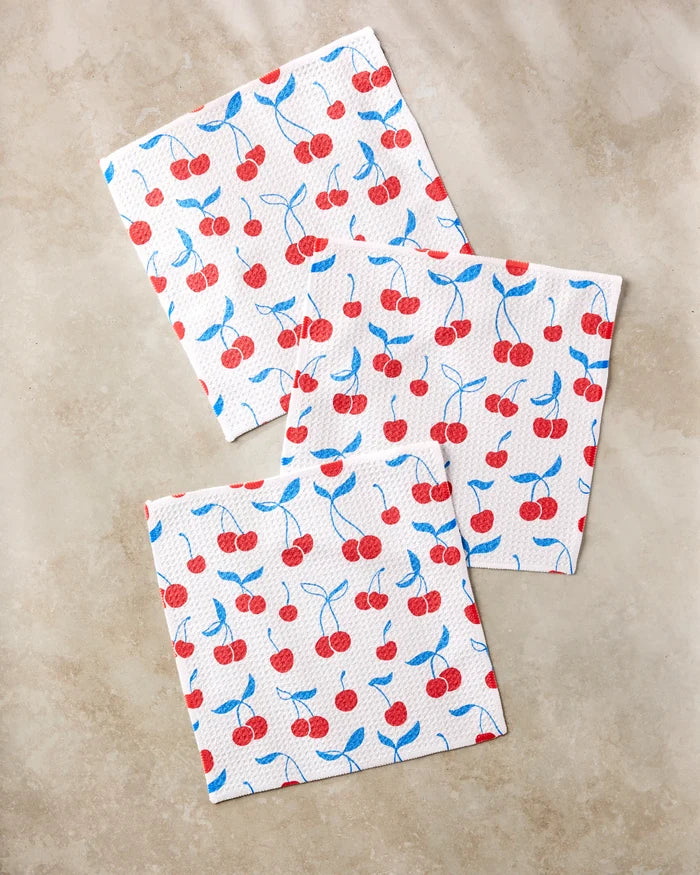Three "Very Cherry" square paper napkins by Kayla Ann, featuring red cherries and blue leaves, are arranged on a beige marble surface for a playful accent beside your recycled dishcloths or kitchen towels.