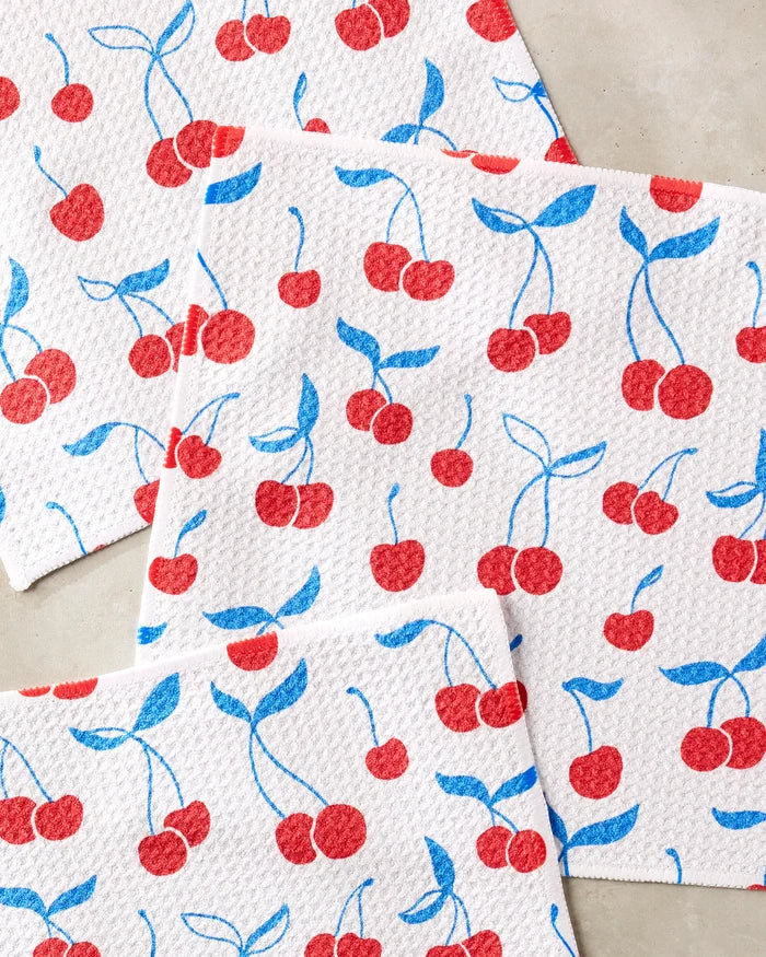 The Kayla Ann Very Cherry set includes four white kitchen towels, each with a repeating red cherry pattern with blue stems and leaves, displayed on a light-colored surface.