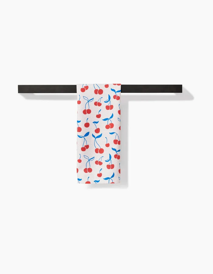 The Kayla Ann Very Cherry Bar Towel, crafted from premium Geoweave fabric, showcases a red cherry and blue stem design and hangs neatly on a horizontal black bar against a plain white background.