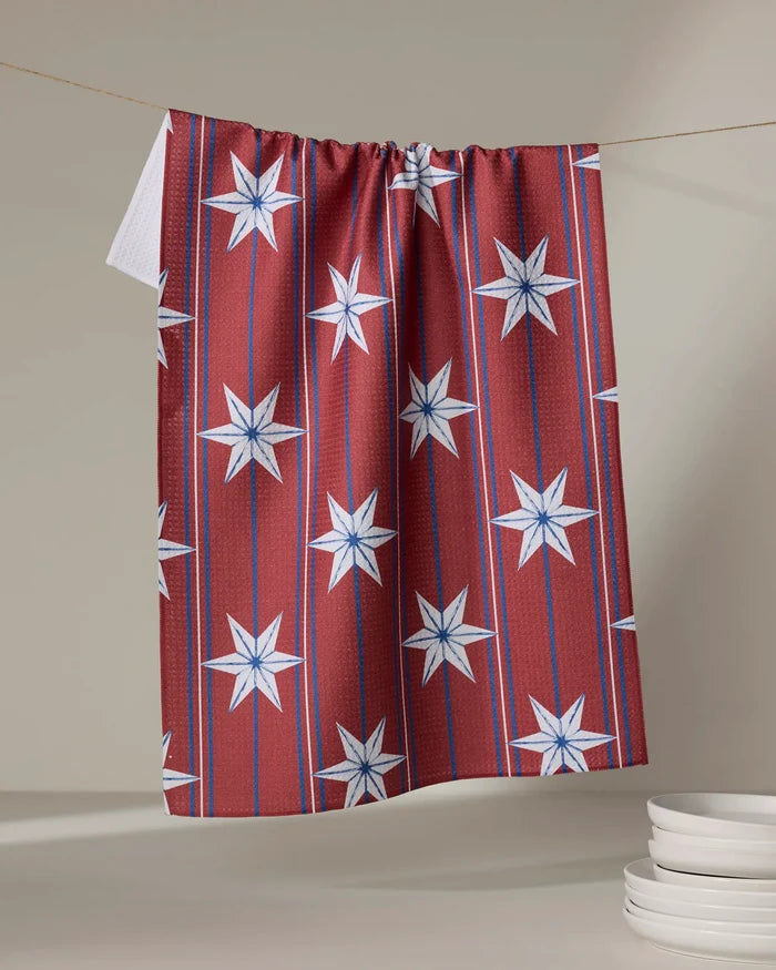 The Ashley Celena Vintage Stars tea towel, featuring blue and white star patterns and vertical stripes, hangs on a clothesline above a stack of white plates. Its quick-drying design adds convenience to your kitchen.