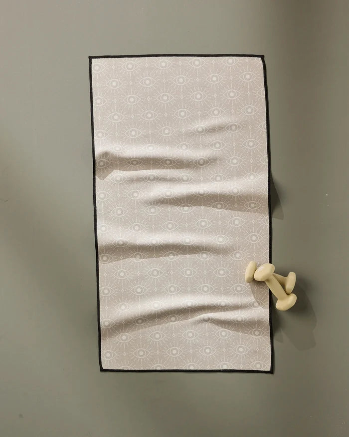 The Geometry Vision towel is a rectangular, light pink fitness towel with a subtle eye pattern. Ultra-absorbent and quick-drying, it hangs on a gray wall from a cream-colored, abstract hook shaped like bent legs.