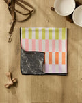 A folded Wag Wise towel by Geometry, featuring a colorful checkered pattern and black underside, sits on a light wood floor near two white pet bowls on a wooden stand, a brown leash, and two dog bones.