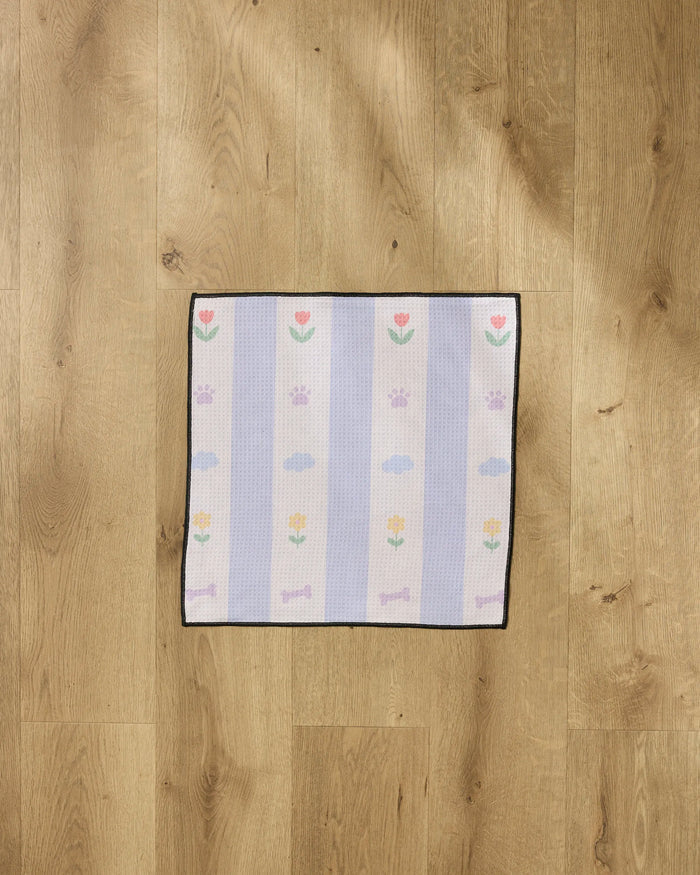 The Wagging Blooms Paw Towel by Letters By Shells is a small rectangular towel with pastel stripes, floral, paw print, bone, and cloud patterns. Made from ultra-absorbent recycled materials, it's shown on a light wooden floor.