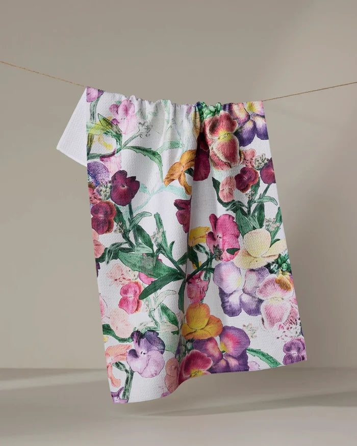 The Wallflower tea towel by Pascale Dilger features ultra-absorbent fabric and a vibrant floral pattern in pink, purple, yellow, and green blooms, shown hanging on a clothesline against a neutral backdrop.