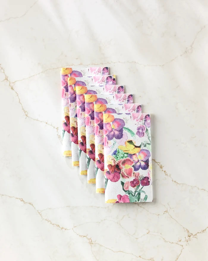 Seven Wallflower chocolate bars by Pascale Dilger, featuring bright floral watercolor packaging, are arranged in a neat diagonal line on a light marble surface, evoking the freshness of reusable paper towels.