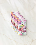 Seven Wallflower chocolate bars by Pascale Dilger, featuring bright floral watercolor packaging, are arranged in a neat diagonal line on a light marble surface, evoking the freshness of reusable paper towels.