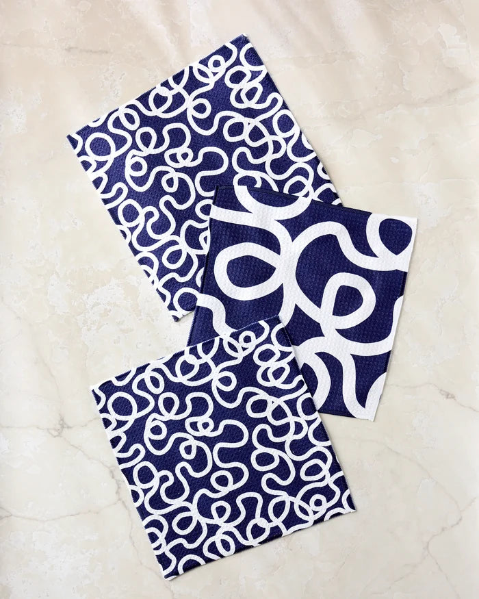 Three “Wander” kitchen towels by Rachel Parker in navy and white, featuring abstract squiggly lines—two with dense patterns and one with larger loops—are arranged on a light marble surface.