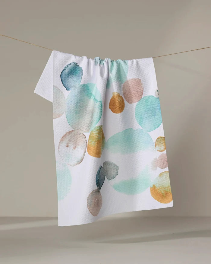 The Taryn Brennan Watercolor Rocks tea towel, made from recycled materials and featuring large teal, gold, peach, and beige watercolor-style circles, hangs on a line against a neutral background.