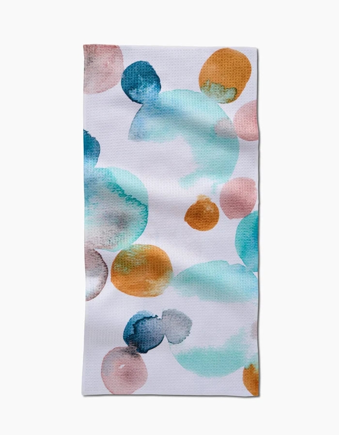 The Taryn Brennan Watercolor Rocks bar towel features a rectangular white base with a vibrant abstract pattern of overlapping blue, teal, orange, and pink circles in watercolor style, crafted from Geoweave fabric.