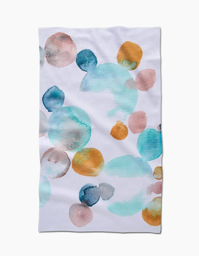 The Watercolor Rocks tea towel by Taryn Brennan features abstract watercolor circles in blue, teal, orange, and peach on white Geoweave fabric, arranged in a scattered, overlapping design.