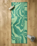 The GEOMETRY Watermelon Vibes green yoga mat with abstract waves is paired with a non-slip towel, a light gray water bottle in the top left, and light blue yoga blocks at the bottom right on a wood floor.