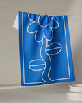 The Wavy Daisy blue tea towel by Beetle Ink Co., made from recycled materials and featuring a white line-drawn flower, hangs on a string above stacked white dishes in a light, minimal setting.