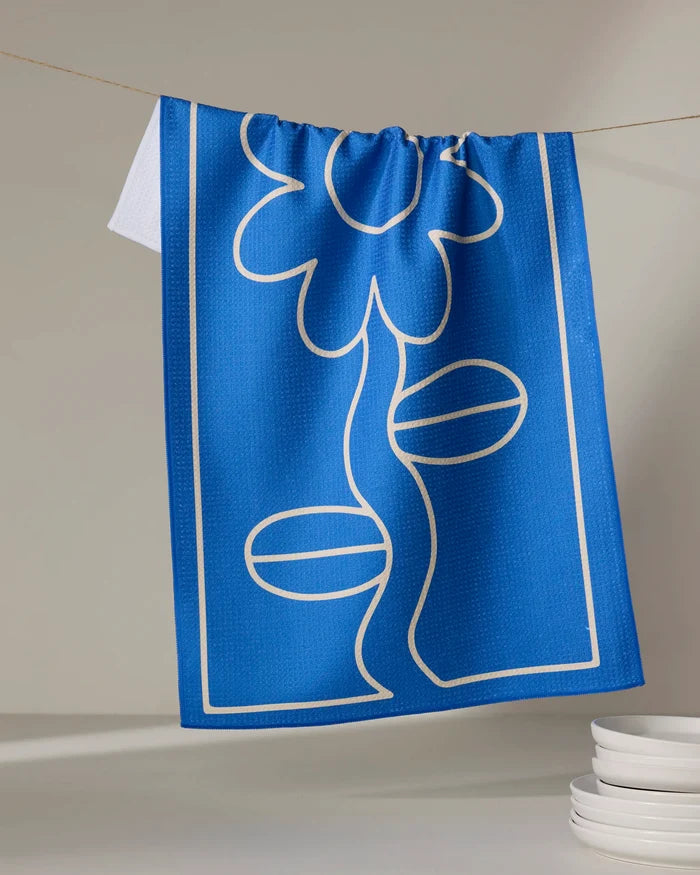 The Wavy Daisy blue tea towel by Beetle Ink Co., made from recycled materials and featuring a white line-drawn flower, hangs on a string above stacked white dishes in a light, minimal setting.