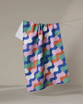 The Claire Iglesias "Wavy Stripes" towel, featuring a vibrant geometric pattern in blue, green, pink, purple, and orange, hangs on a clothesline against a neutral background.