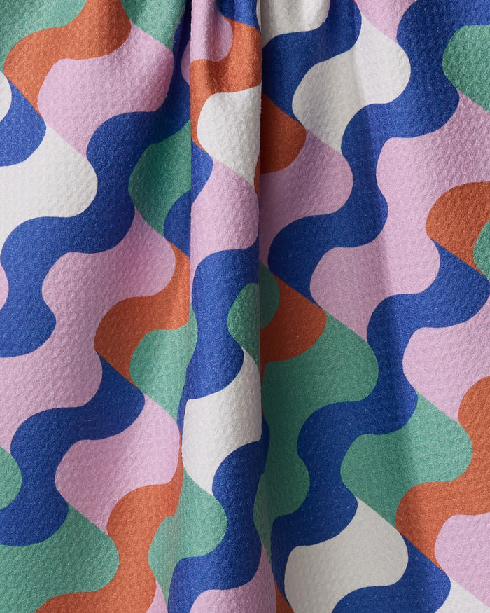 The Wavy Stripes kitchen towel by Claire Iglesias features a colorful wavy pattern in blue, green, orange, white, and lavender on textured recycled fabric with a vertical center fold.