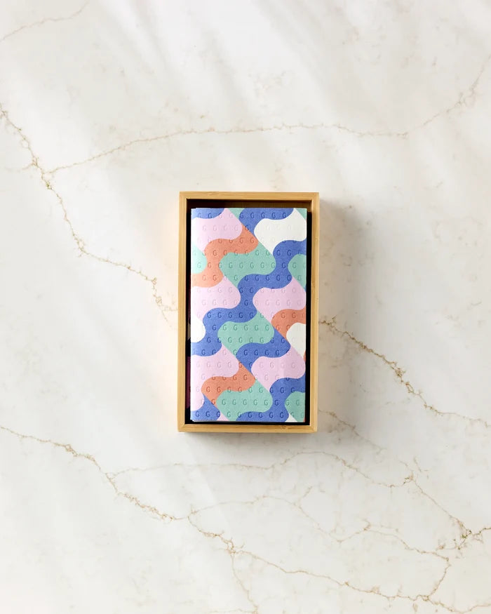 The Claire Iglesias Wavy Stripes rectangular box of eco-friendly paper towels, featuring a pastel abstract pattern in blue, pink, purple, and peach, sits centered on a light marble surface.