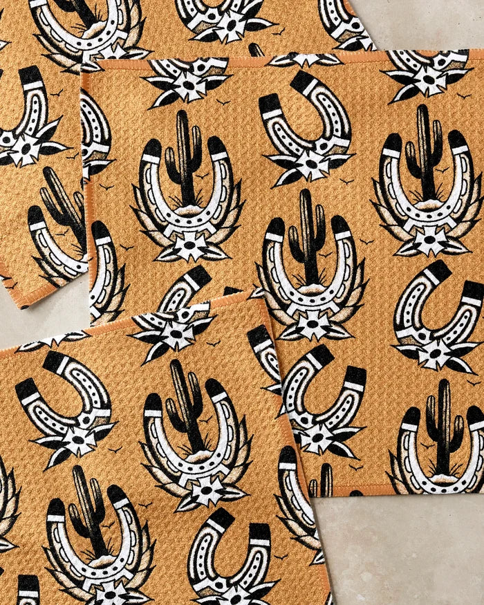 The Cole Strem Western Horseshoes set features patterned dishcloths with black and white horseshoes, cacti, and stars on a textured tan background. Multiple super-absorbent towels are layered and slightly overlapping.