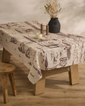 On a wooden table rests Geometrys Western Scene lightweight, stain-resistant tablecloth with nature designs. The setting includes a black dried-flower vase, two salt and pepper shakers, white cup and saucer, and a wooden stool on wood flooring.