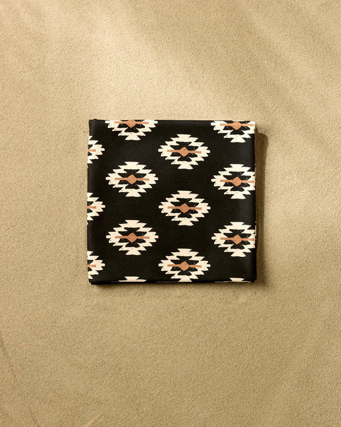 The Hufton Studio Western Tile is a folded beach blanket made from recycled materials, showcasing a black base with a white and brown southwestern geometric pattern, displayed on a beige surface.