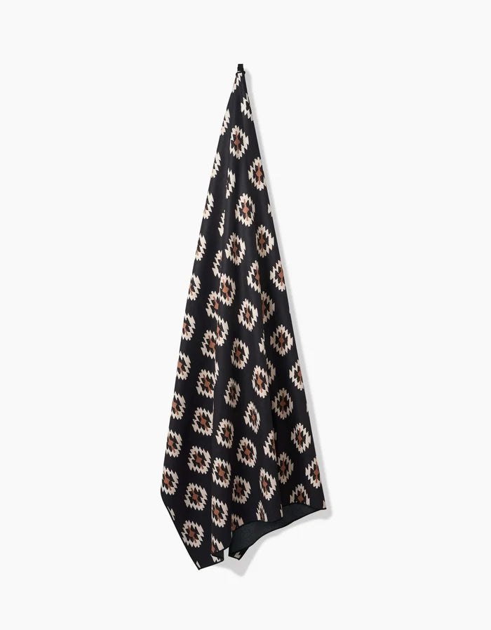 A Western Tile Beach Blanket by Hufton Studio, made from recycled materials with a black base and geometric white, brown, and tan pattern, hangs against a plain white background.
