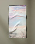 “When I Close My Eyes” by Stefanie Bales is a rectangular, ultra-absorbent cloth featuring a printed calm sea and pastel sky with clouds, bordered in black and displayed against a plain gray wall.