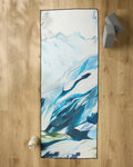 The "Where He Laid To Go" non-slip yoga towel by Kelly Peters, featuring a blue and white abstract mountain design, is on a wooden floor. A water bottle sits bottom left, with two gray yoga blocks stacked at the top right.