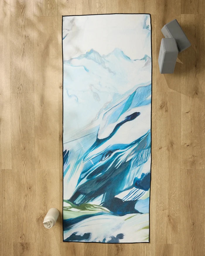 The "Where He Laid To Go" non-slip yoga towel by Kelly Peters, featuring a blue and white abstract mountain design, is on a wooden floor. A water bottle sits bottom left, with two gray yoga blocks stacked at the top right.