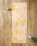 The GEOMETRY Whimsy yoga mat, featuring a pastel pink and yellow abstract pattern, is laid on a wooden floor with a non-slip towel. Alongside it are a white water bottle and gray yoga blocks.