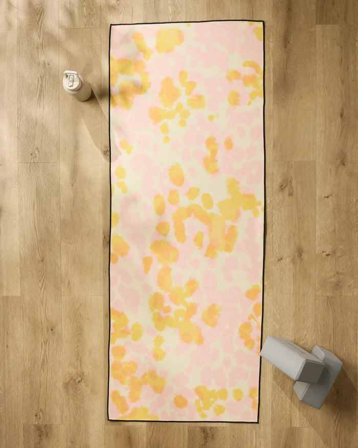 The GEOMETRY Whimsy yoga mat, featuring a pastel pink and yellow abstract pattern, is laid on a wooden floor with a non-slip towel. Alongside it are a white water bottle and gray yoga blocks.