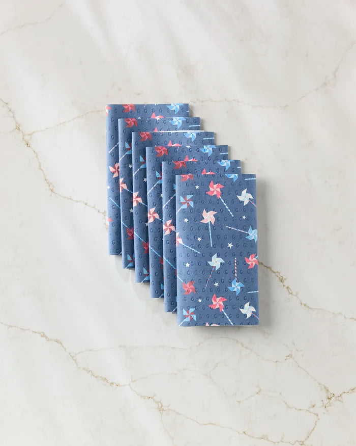 Six Whirlwind Pins blue napkins by Rebecca Bobko, featuring red and light blue pinwheels, are neatly arranged on a light marble surface—an eco-friendly alternative to disposable paper towels.