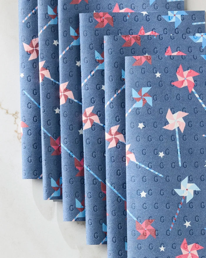 Six folded Whirlwind Pins cloth napkins by Rebecca Bobko, featuring red, white, and blue pinwheels, small stars, and G’s—an eco-friendly set arranged on a white marble surface.