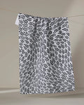 The Rachel Parker White Leaf Swirl tea towel, featuring a black leafy pattern on white fabric, hangs from a thin line against a beige background, its slightly wrinkled cloth appearing to float in mid-air.