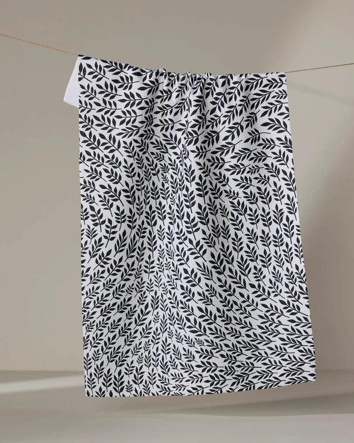 The Rachel Parker White Leaf Swirl tea towel, featuring a black leafy pattern on white fabric, hangs from a thin line against a beige background, its slightly wrinkled cloth appearing to float in mid-air.