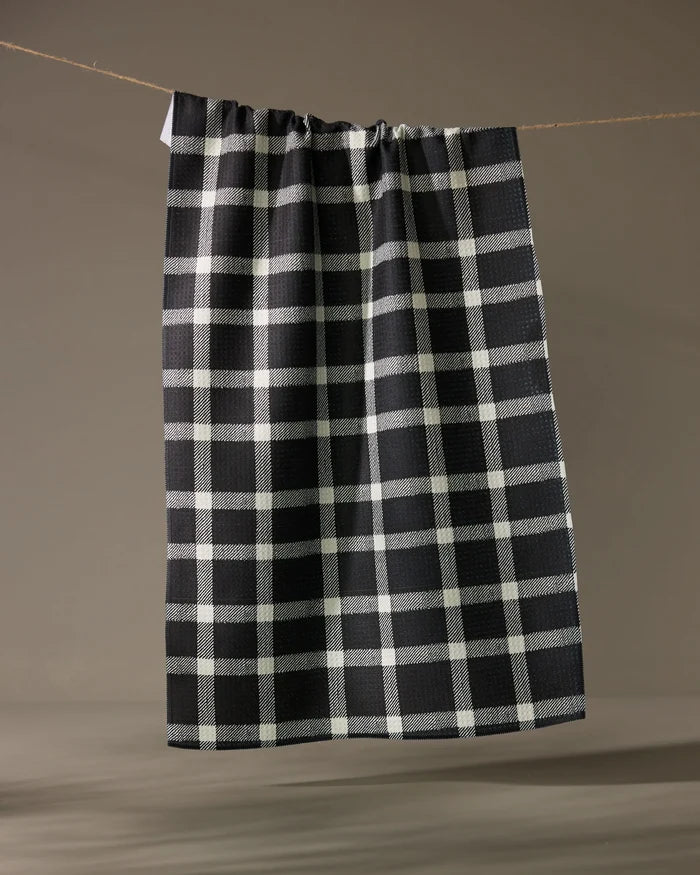 The Geometry Wicklow Black kitchen towel, featuring a black and white checkered pattern, hangs on a clothesline against a simple, neutral background.