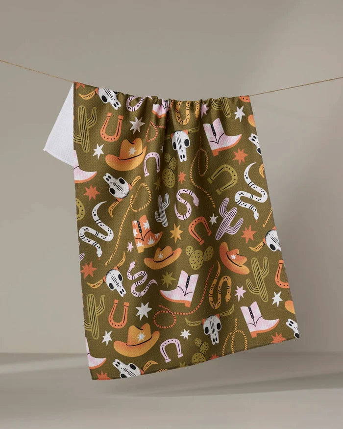 The "Wild Cowboy" tea towel by Megan Roy, made from recycled materials, features a colorful western pattern with cowboy hats, boots, cacti, horseshoes, snakes, stars, and bull skulls. It hangs on a line against a plain background.