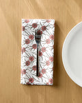 A silver fork rests on a Geometry Wild Dahlia reusable cloth napkin with pink flowers and green leaves, placed on a wooden table next to a white plate.
