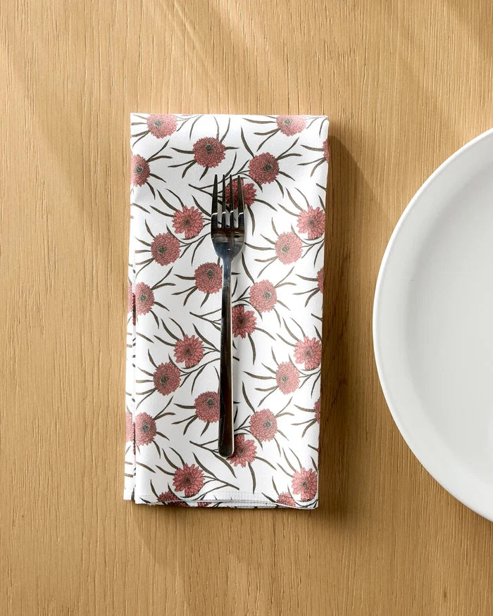 A silver fork rests on a Geometry Wild Dahlia reusable cloth napkin with pink flowers and green leaves, placed on a wooden table next to a white plate.