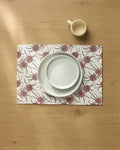 A white mug and two white plates are stacked on the Geometry Wild Dahlia recycled placemat, featuring red and purple flowers, all arranged on a light wood table.