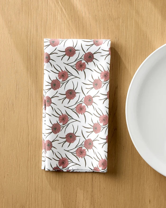 The Geometry Wild Dahlia white reusable cloth napkin, featuring a pink and green floral pattern, is neatly folded on a wooden table beside a plain white plate.