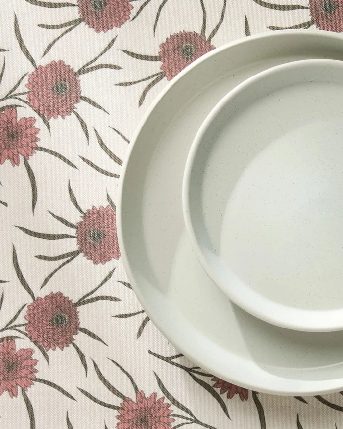 A smaller light gray ceramic plate nests in a larger one atop Geometrys Wild Dahlia placemats, which feature red flowers and green stems, creating a simple and elegant table setting.