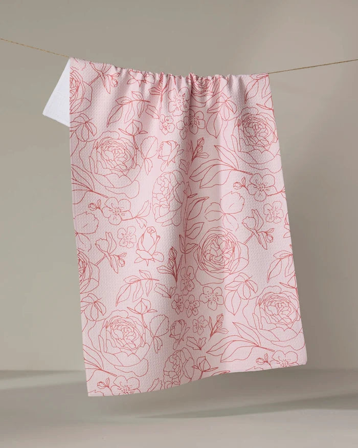 The Wild Roses tea towel by Janna Sue Design features a pink, absorbent fabric with a red floral line-drawing pattern, hanging on a clothesline against a plain, light background.