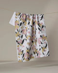 The Toboggan Avenue Wilderness Floral quick-drying towel, featuring a pink, yellow, and black floral design, hangs on a line against a simple beige background.