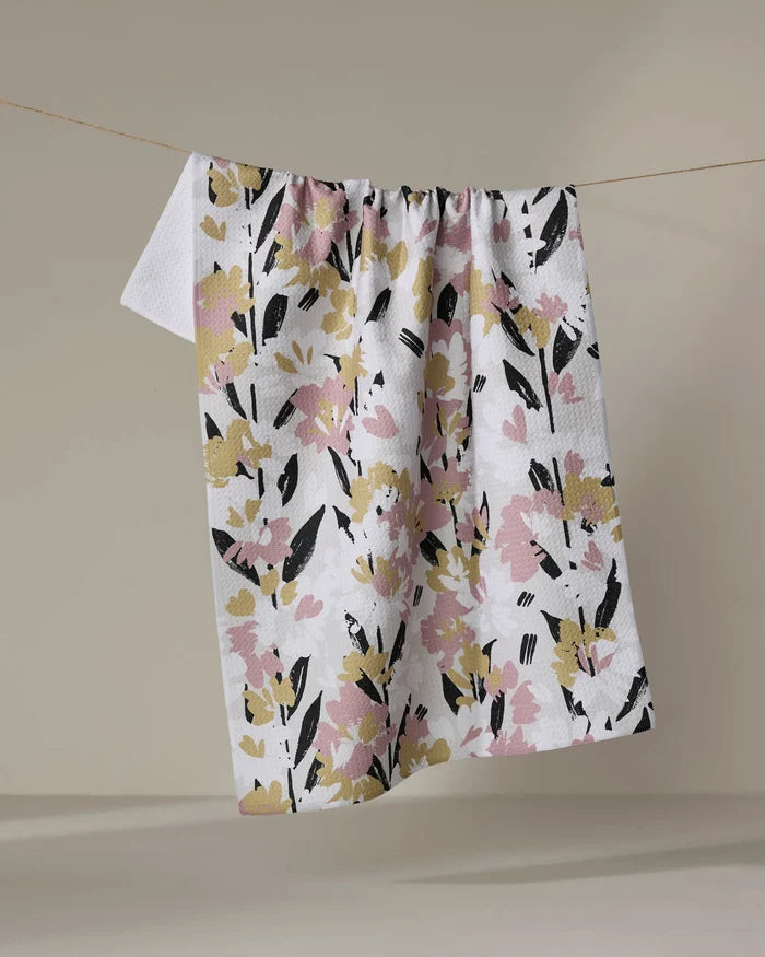 The Toboggan Avenue Wilderness Floral quick-drying towel, featuring a pink, yellow, and black floral design, hangs on a line against a simple beige background.