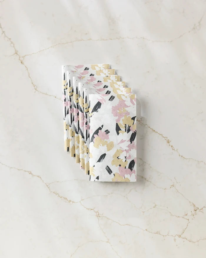 A stack of Toboggan Avenue Wilderness Floral notebooks with pink, yellow, and black accents arranged on marble next to eco-friendly paper towels.