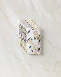 A stack of Toboggan Avenue Wilderness Floral notebooks with pink, yellow, and black accents arranged on marble next to eco-friendly paper towels.