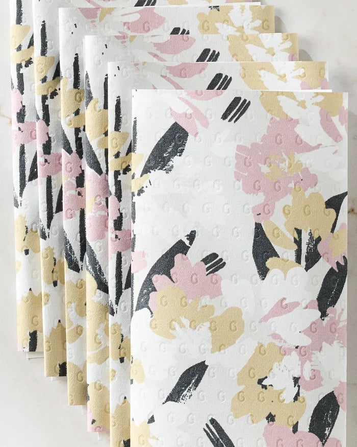 Eight eco-friendly Wilderness Floral paper towels by Toboggan Avenue, featuring pink, yellow, black, and white floral designs, are folded and fanned out on a white surface.