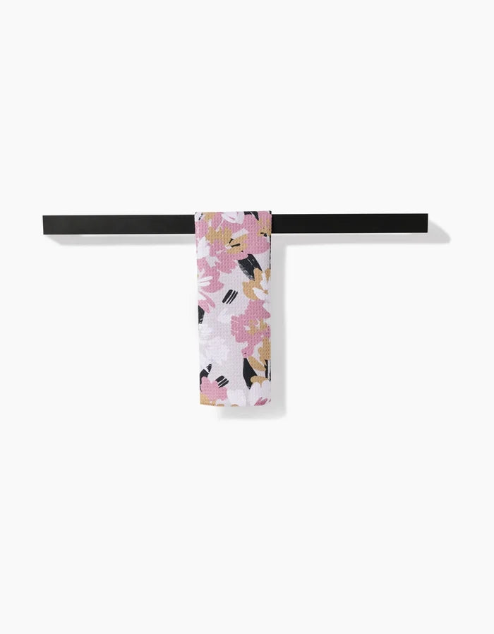 The Toboggan Avenue Wilderness Floral Bar Towel, made from recycled materials in pink, white, black, and mustard florals, is neatly draped over a minimalist black towel bar against a plain white background.